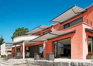 Moderne woning met meerdere zonneschermn en een terras, omringd door groene bomen en een blauwe lucht. 