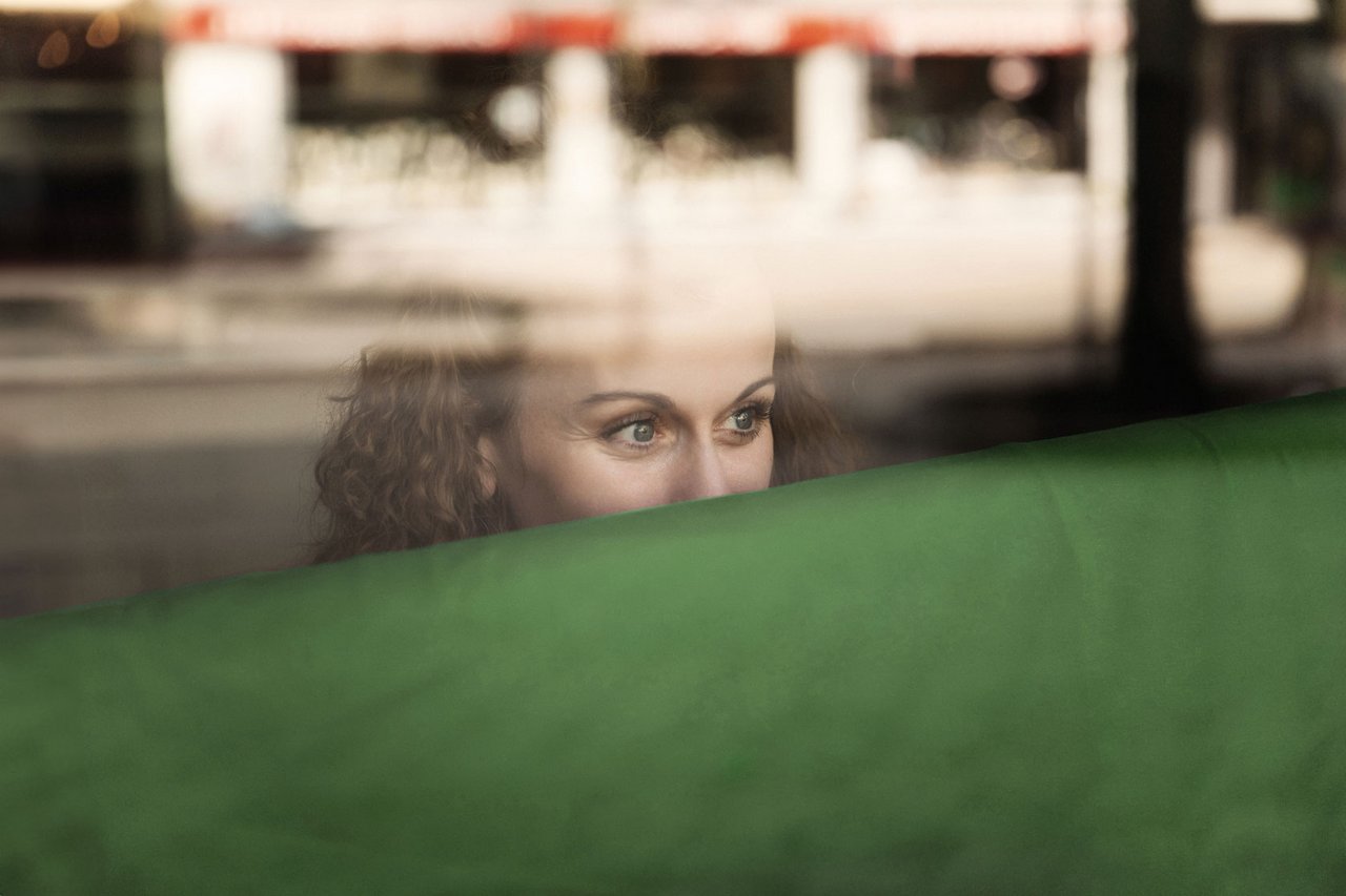 Eine Frau mit Locken blickt neugierig durch ein Fenster, das eine grüne Fläche reflektiert.
