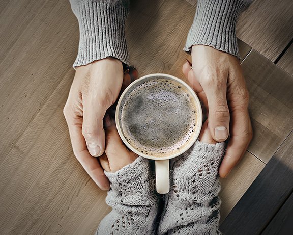 Twee handen houden een koffiekopje vast, omringd door een houten tafel. Twee handen houden een koffiekopje vast, omringd door een houten tafel.