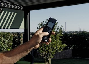 Vinger die een afstandsbediening vasthoudt met een uitzicht op een groene tuin en een blauwe lucht. 
