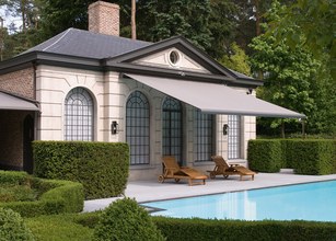 Elegant huis met zonnescherm boven het terras, omringd door groene hagen en een zwembad. 