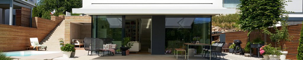 Moderne woning met grote glaspartijen en een verzorgde terrassensituatie. Groene planten en houten elementen aanwezig.