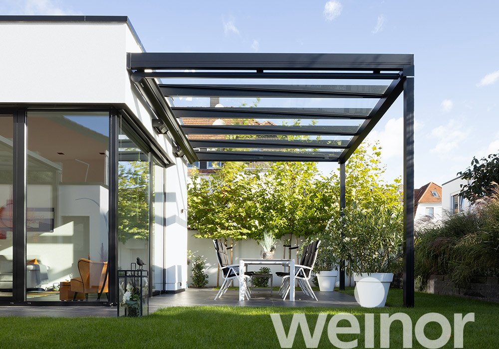 Een moderne overkapping met glazen schuifwanden en een terras met witte meubels en groene planten. 