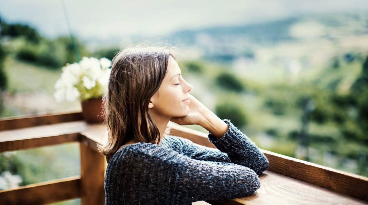 Vrouw met een serene blik, zittend op een houten balkon met achtergrond van een groene natuur.