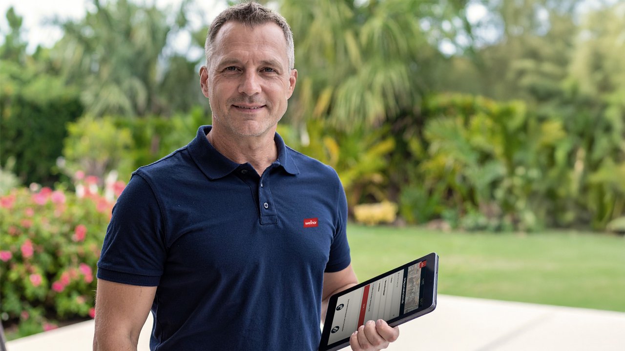 Ein Mann in einem dunkelblauen Shirt hält ein Tablet und steht in einem blühenden Garten.