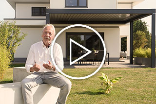 Man met een man die buiten zit met een huis en een overkapping op de achtergrond. Groen gras en planten zichtbaar.