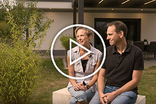 Een vrouw en een man zitten op een muurtje in een tuin met groene planten en een moderne woning op de achtergrond.