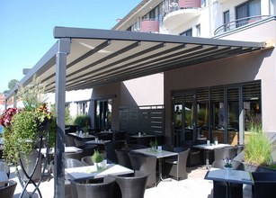 Een terras met een moderne overkapping, voorzien van tafels en stoelen. Groene planten zorgen voor een sfeervolle uitstraling. 