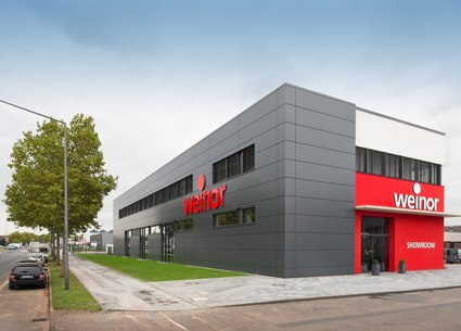 Modern gebouw met een grijze gevel en rode accenten; prominent logo van weinor aan de voorkant.