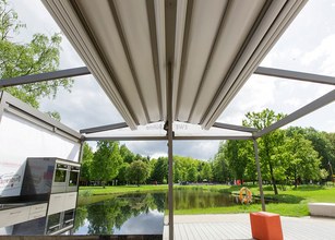 Terrassensituatie met een overkapping, uitzicht op een vijver en groene boompartijen. 