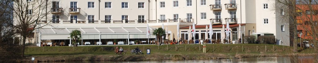 Een lichtgekleurde, moderne gebouw met een terras en groen gras aan een waterkant. Een lichtgekleurde, moderne gebouw met een terras en groen gras aan een waterkant.
