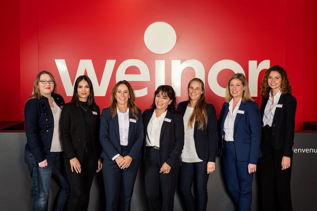 Groep vrouwen in zakelijke kleding voor een rode achtergrond met een merknaam.